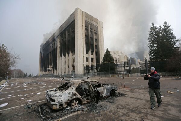 Vyras fotografuoja apdegusį automobilį priešais savivaldybės pastatą, kuris buvo padegtas per protestus, kilusius dėl degalų kainų didėjimo Almatoje, Kazachstane 2022 metų sausio 6 dieną. - Sputnik Lietuva