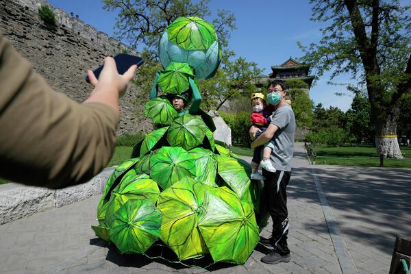 Pekine minint Žemės dieną, vyras ir vaikas, dėvintys kaukes, pozuoja nuotraukoms šalia kinų menininkės Kong Ning su naujausiu kūriniu &quot;Protect Earth with Banana Leaf Umbrellas&quot;. - Sputnik Lietuva