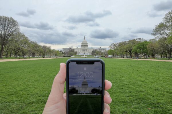 Показывающие полдень часы в мобильном телефоне напротив Капитолия в Вашингтоне в период пандемии коронавируса - Sputnik Литва
