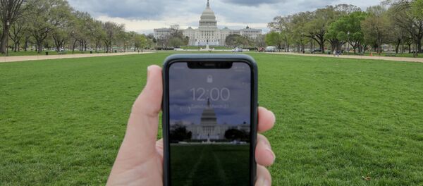 Показывающие полдень часы в мобильном телефоне напротив Капитолия в Вашингтоне в период пандемии коронавируса Показывающие полдень часы в мобильном телефоне напротив Капитолия в Вашингтоне в период пандемии коронавируса - Sputnik Литва